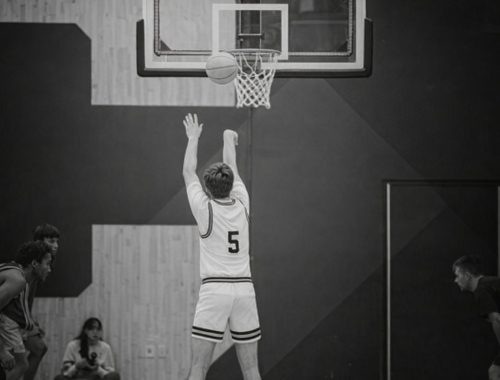 Svartvit bild på basketspelare som kastar boll mot korg i en hall.