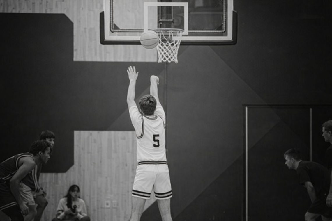 Svartvit bild på basketspelare som kastar boll mot korg i en hall.