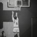 Svartvit bild på basketspelare som kastar boll mot korg i en hall.