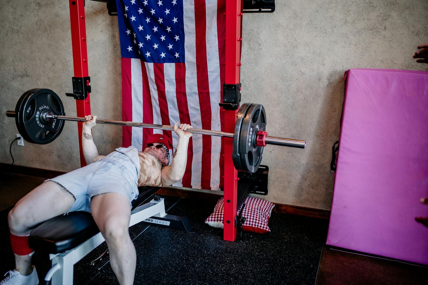 En person utför bänkpress med skivstång i ett hemmagym med en amerikansk flagga.